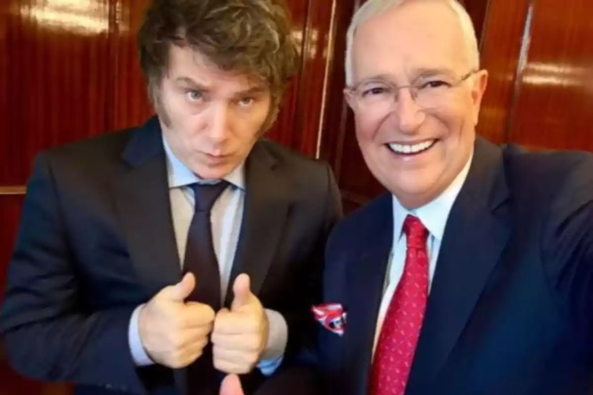 Two men in suits pose for a photo, one of them giving a thumbs-up gesture while the other smiles.