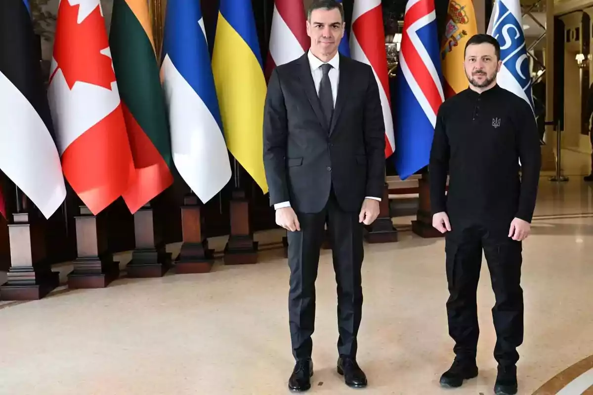 Dos personas de pie frente a una fila de banderas de diferentes países en un entorno formal.