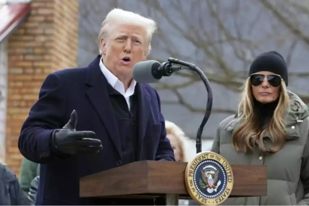 Un hombre hablando en un podio al aire libre con un sello presidencial, acompañado de una mujer con gafas de sol y gorro.