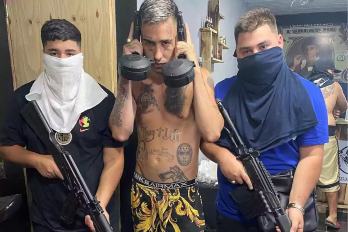 Tres personas posan con armas de fuego, dos de ellas con el rostro cubierto y la persona del centro sin camisa mostrando tatuajes.