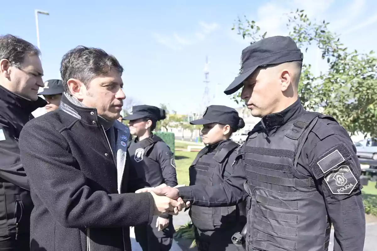 Un hombre con abrigo oscuro estrecha la mano de un oficial de policía uniformado mientras otros oficiales observan en un entorno al aire libre.