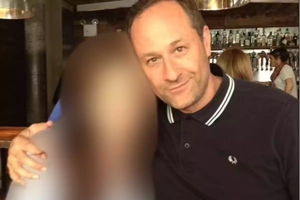 Un hombre con camisa oscura abraza a una persona cuya imagen está borrosa en un bar.