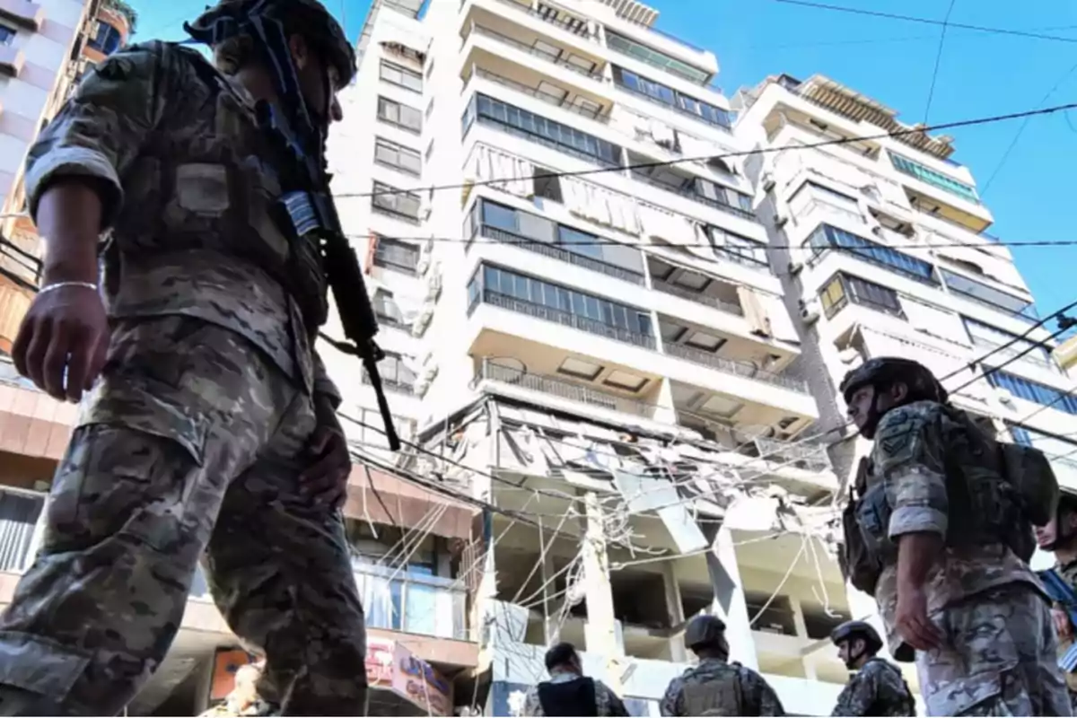 Soldados con uniforme militar y cascos patrullan frente a un edificio alto con ventanas y balcones, mientras cables eléctricos cuelgan en el aire.