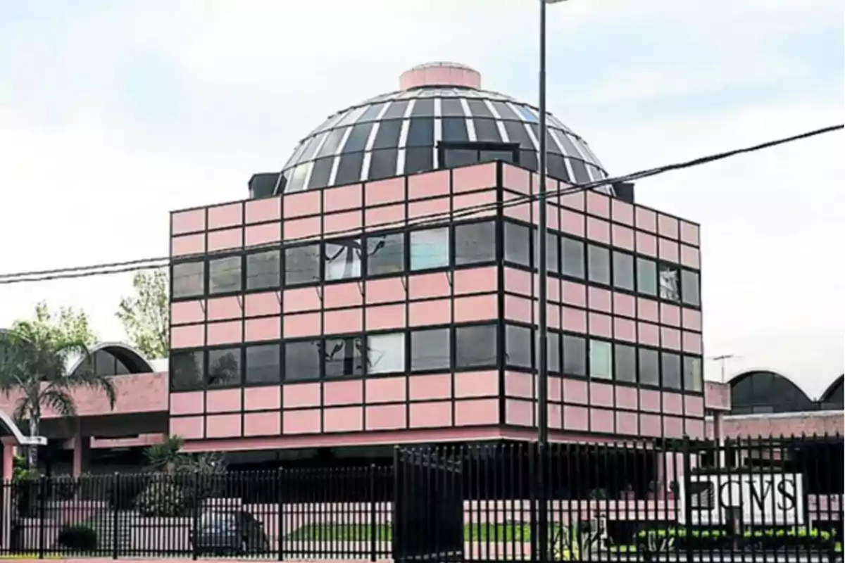 Edificio de oficinas con fachada de vidrio y estructura rosa, coronado por una cúpula de cristal, rodeado por una cerca negra.