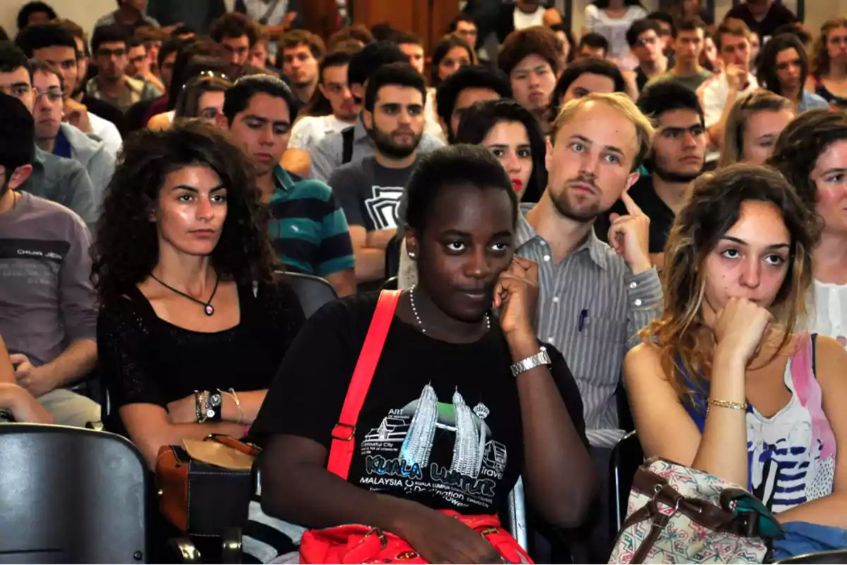 Un grupo de personas sentadas en una conferencia, algunas parecen estar aburridas o distraídas.