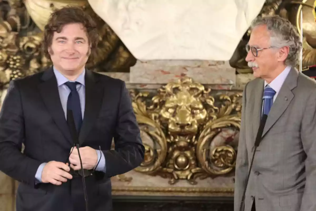 Dos hombres de traje en un evento formal con un fondo decorativo.