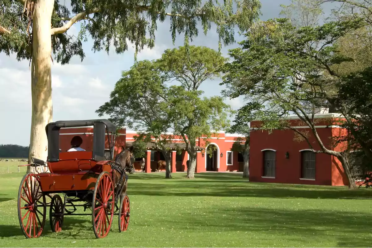 Carruaje tirado por caballo frente a una casa de campo roja rodeada de árboles y césped verde.