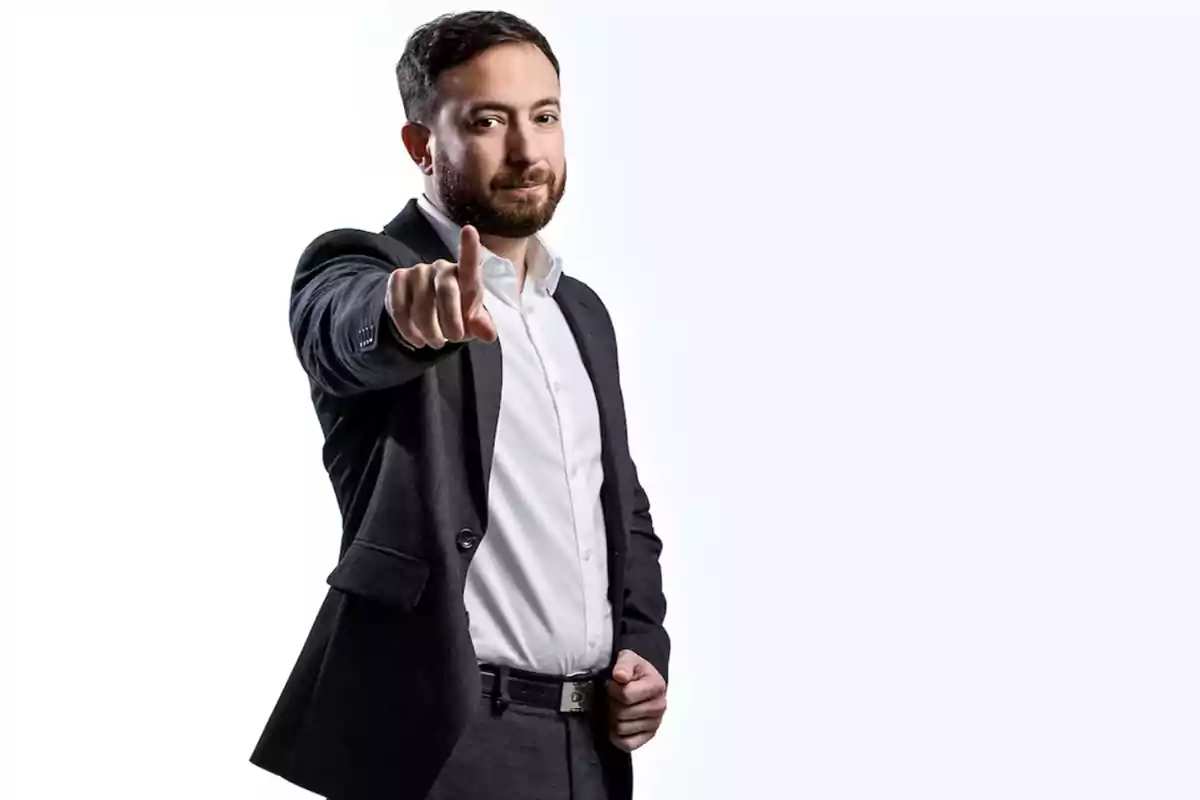 Hombre con barba y traje oscuro señalando hacia el frente con expresión seria.