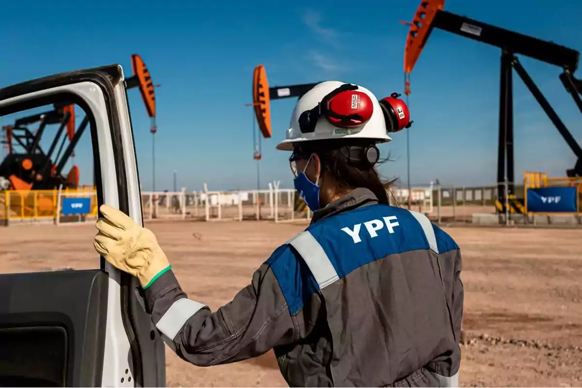 Persona con uniforme de YPF y casco de seguridad observando bombas de extracción de petróleo en un campo.