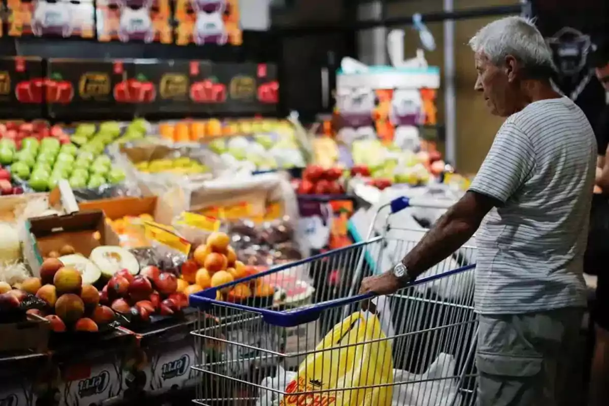 Un hombre mayor con un carrito de compras observa frutas y verduras en un supermercado.