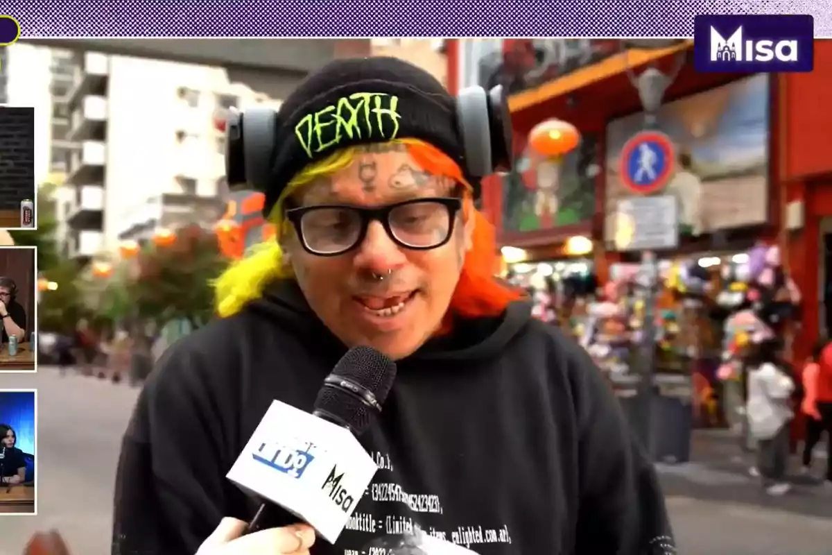 Una persona con cabello de colores y tatuajes en la cara lleva una gorra negra con la palabra "DEATH" mientras sostiene un micrófono en una calle concurrida.