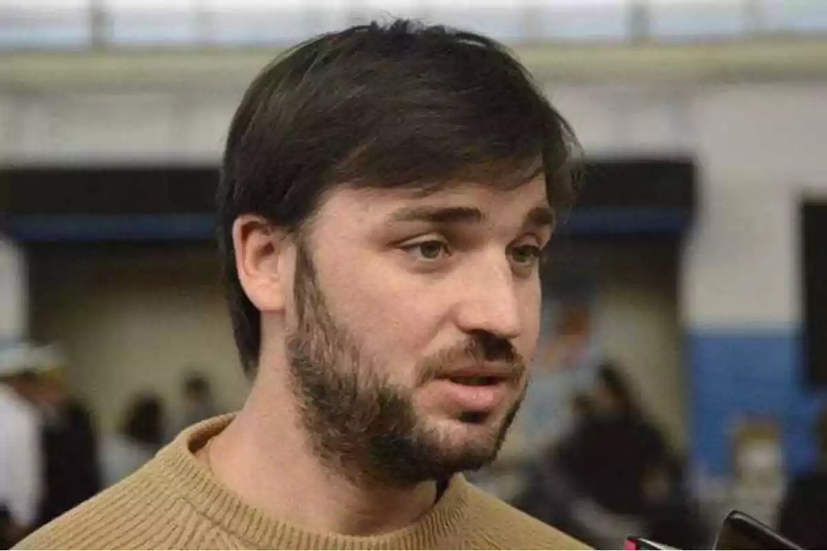 Un hombre con barba y cabello oscuro hablando en un entorno interior.