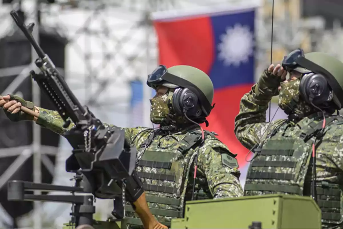 Soldados con uniforme de camuflaje y equipo táctico observan y señalan mientras están junto a una ametralladora, con una bandera de fondo.