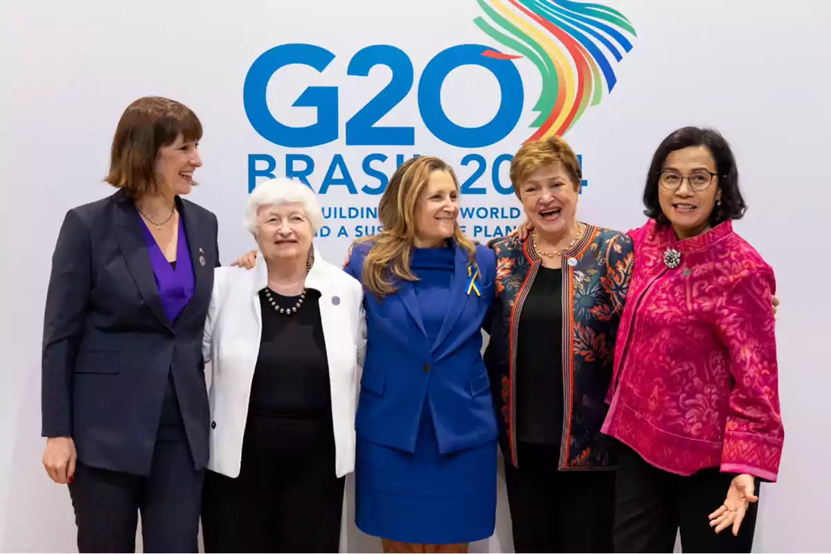 Cinco mujeres posan juntas sonriendo frente a un cartel del G20 en Brasil 2024.