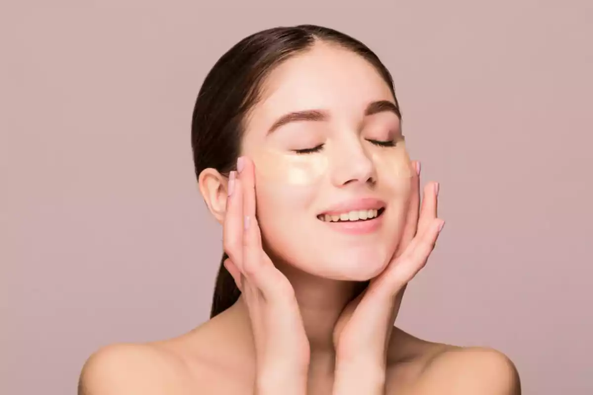 Mujer joven con parches de gel debajo de los ojos sonriendo con los ojos cerrados y las manos en las mejillas.