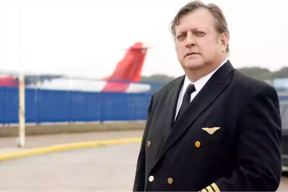 Un hombre con uniforme de piloto de pie en un aeropuerto con un avión en el fondo.