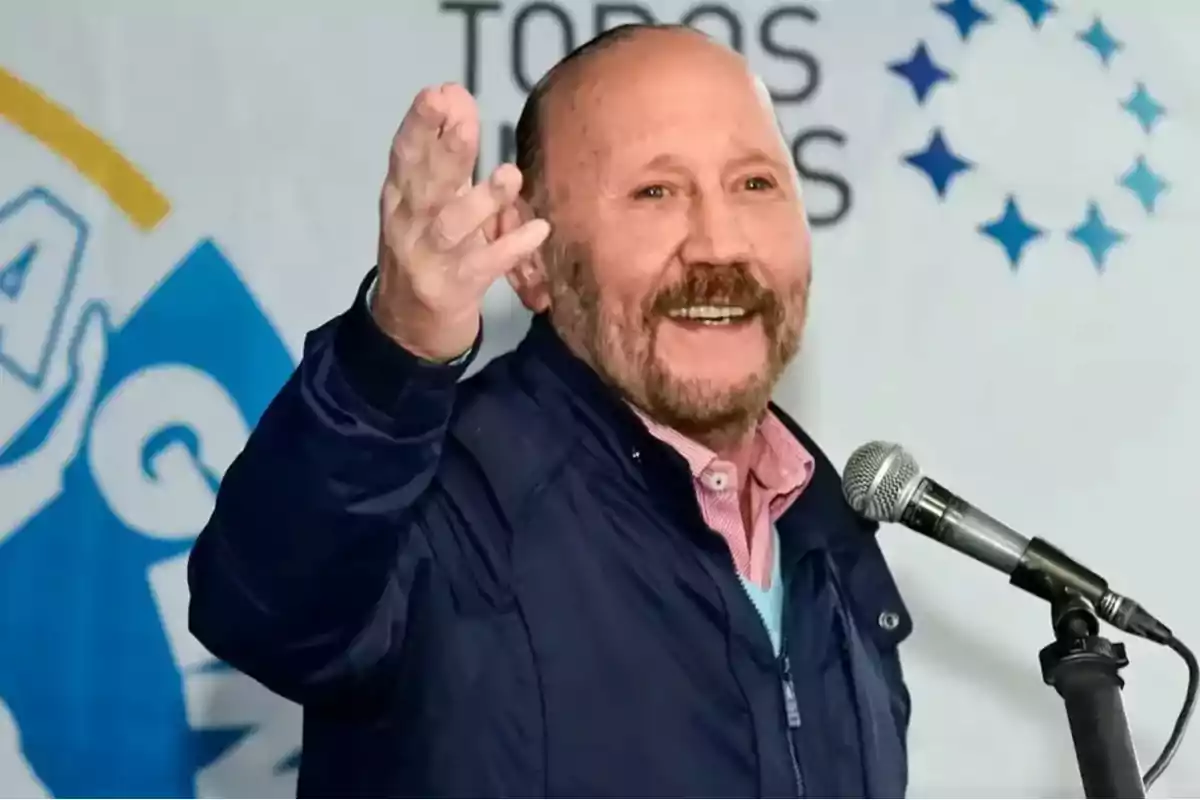 Un hombre sonriente con barba y bigote habla en un micrófono durante un evento, con un fondo que incluye logotipos y texto.