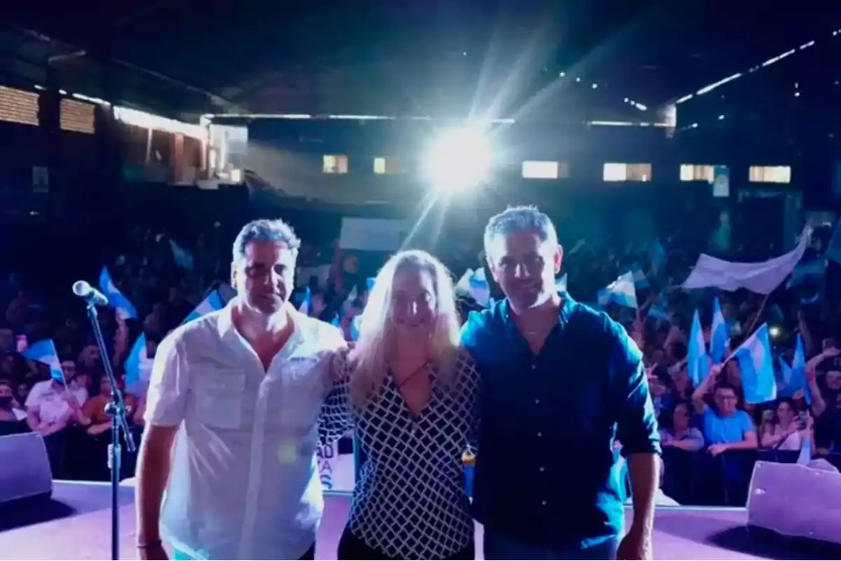 Tres personas posan en un escenario frente a una multitud con banderas en un evento bajo techo.