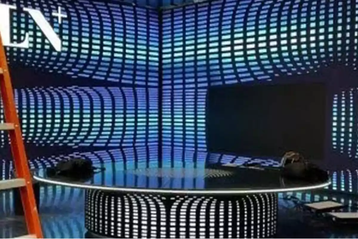 Un estudio de televisión con un diseño futurista en tonos azules y negros, una mesa redonda iluminada y una escalera a la izquierda.