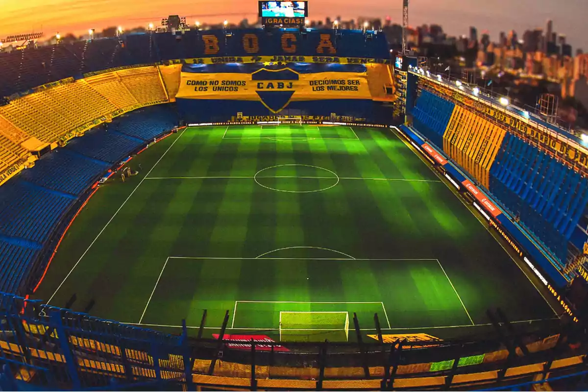 Vista aérea de un estadio de fútbol con gradas azules y amarillas y un campo de juego iluminado al atardecer.