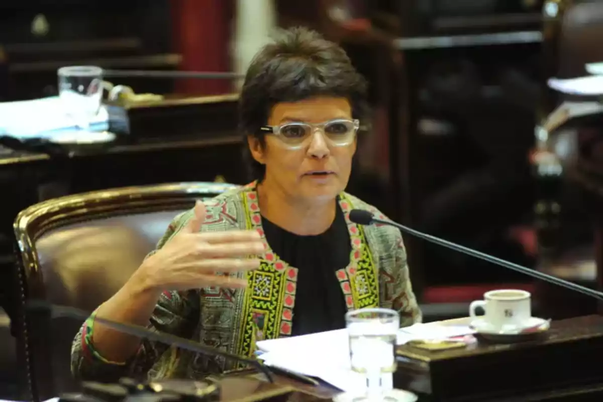 Mujer con gafas hablando en un entorno formal con micrófono y documentos frente a ella.