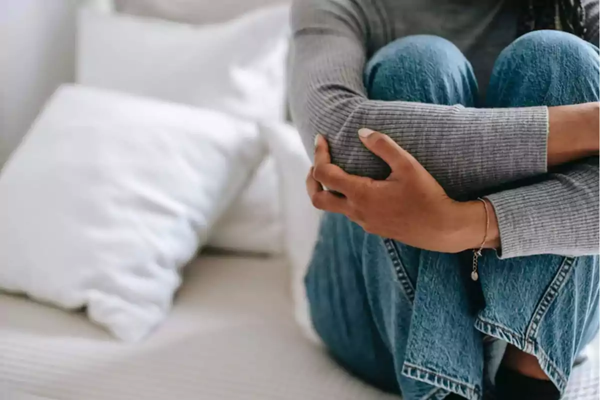 Persona sentada en una cama con las piernas cruzadas, vistiendo un suéter gris y jeans azules.