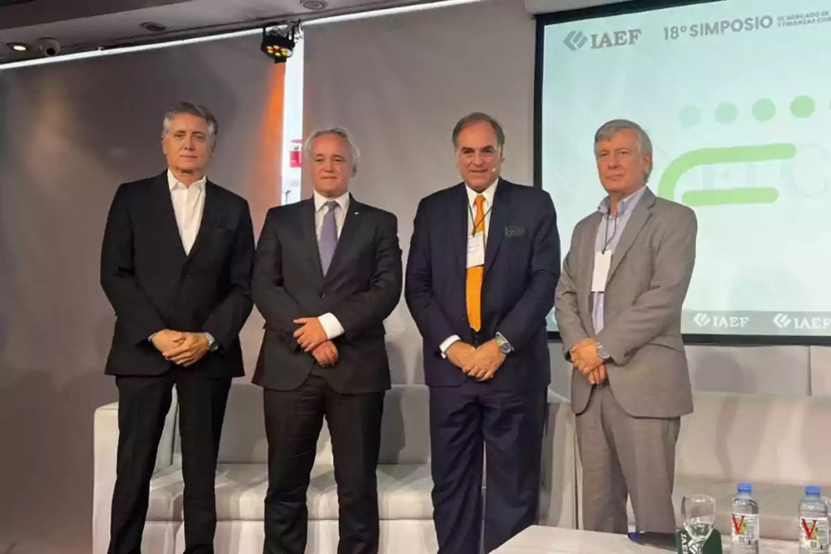 Cuatro hombres de pie en un escenario durante un simposio del IAEF, con un proyector de fondo mostrando el logo del evento.