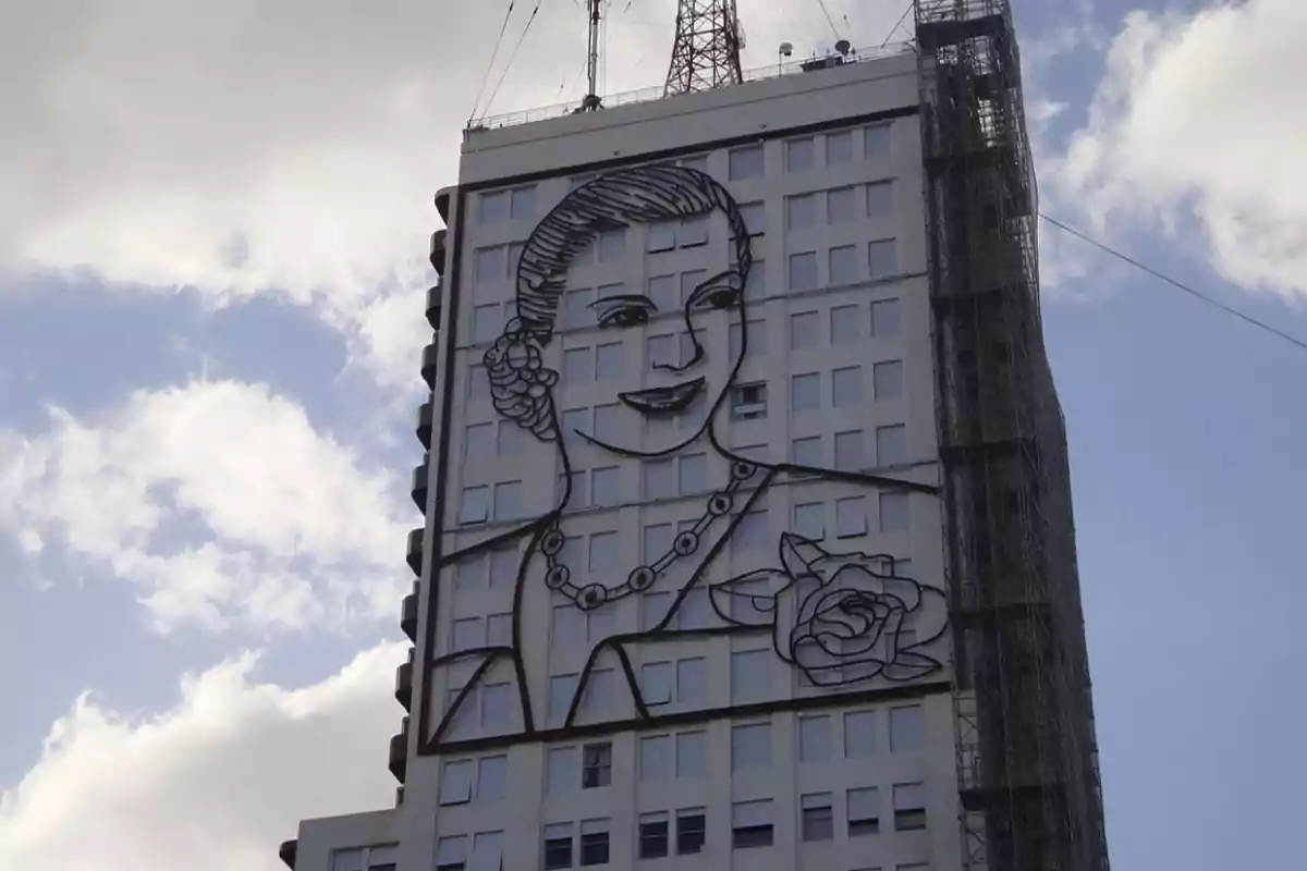 Edificio con un gran mural de una mujer sonriente con un peinado recogido y un collar, sosteniendo una flor, en un día parcialmente nublado.