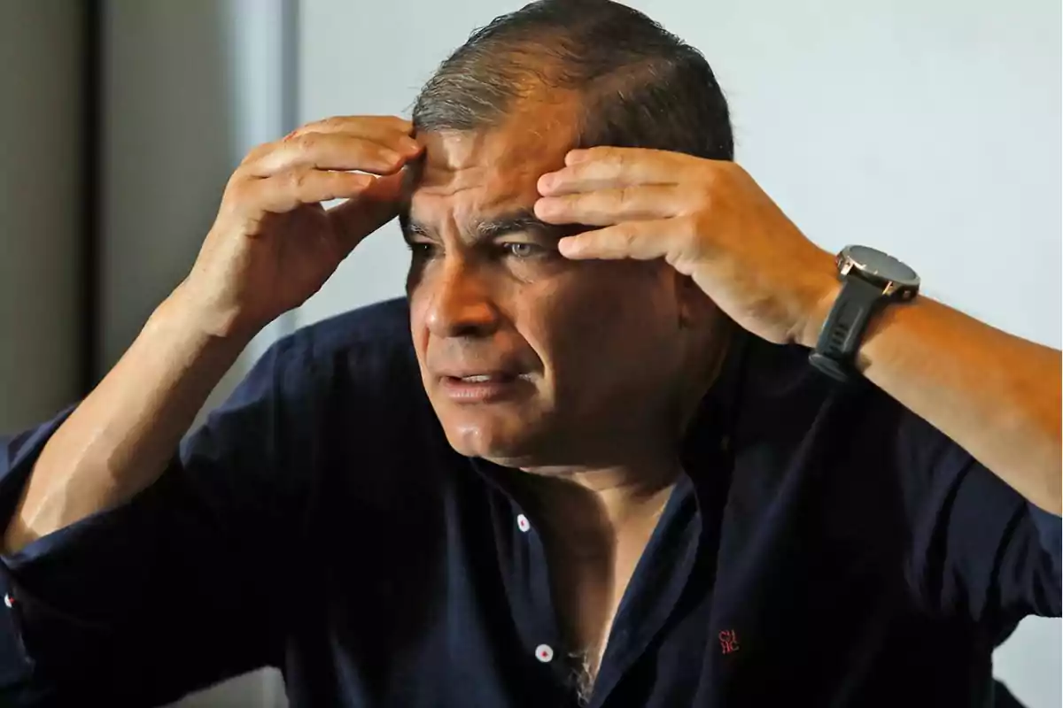 Hombre con camisa oscura y reloj en la muñeca, tocándose la frente con ambas manos.