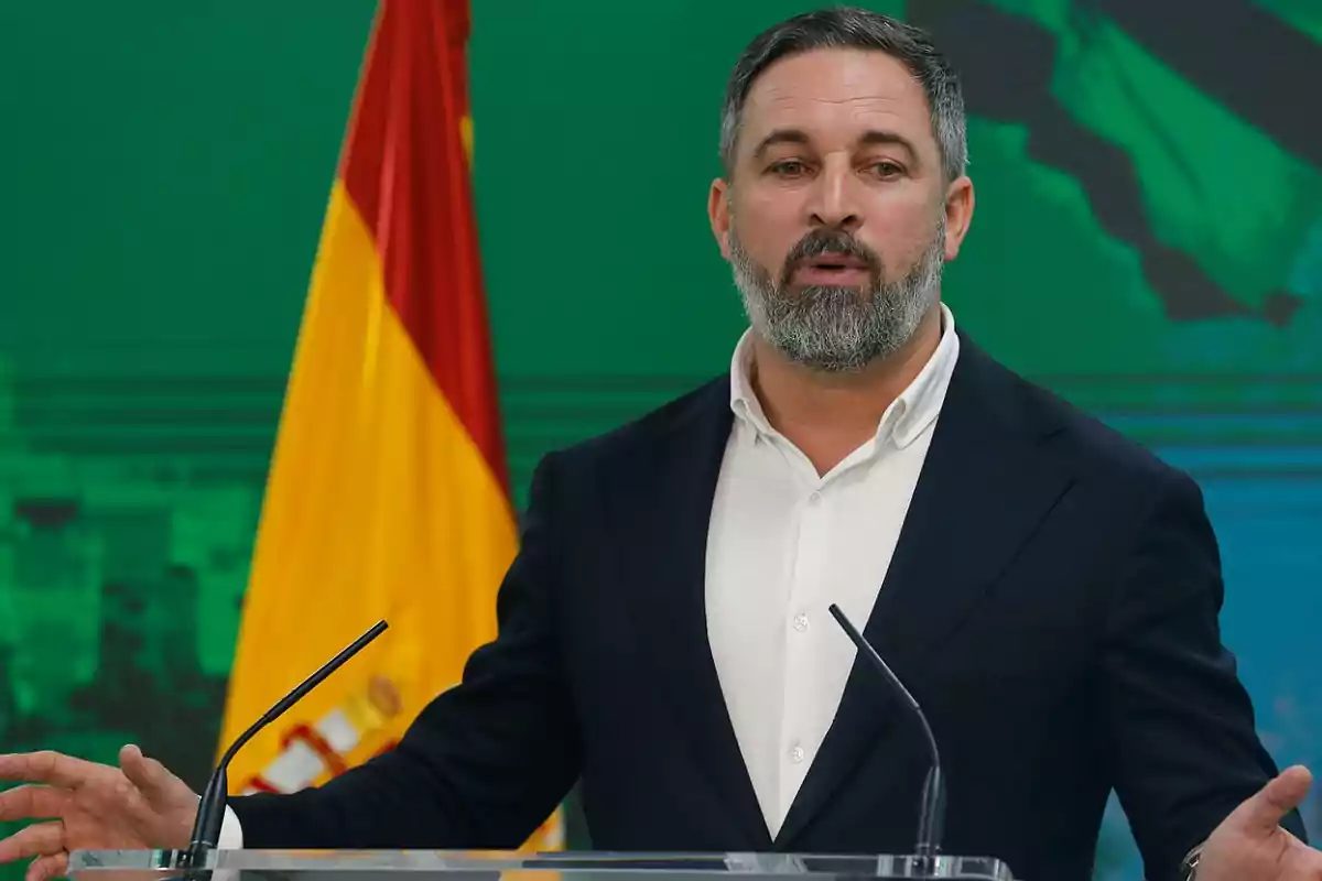 Un hombre de pie frente a un micrófono con una bandera española de fondo.