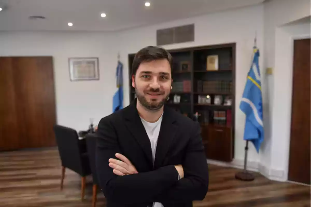 Hombre de pie con los brazos cruzados en una oficina con banderas y estanterías al fondo.