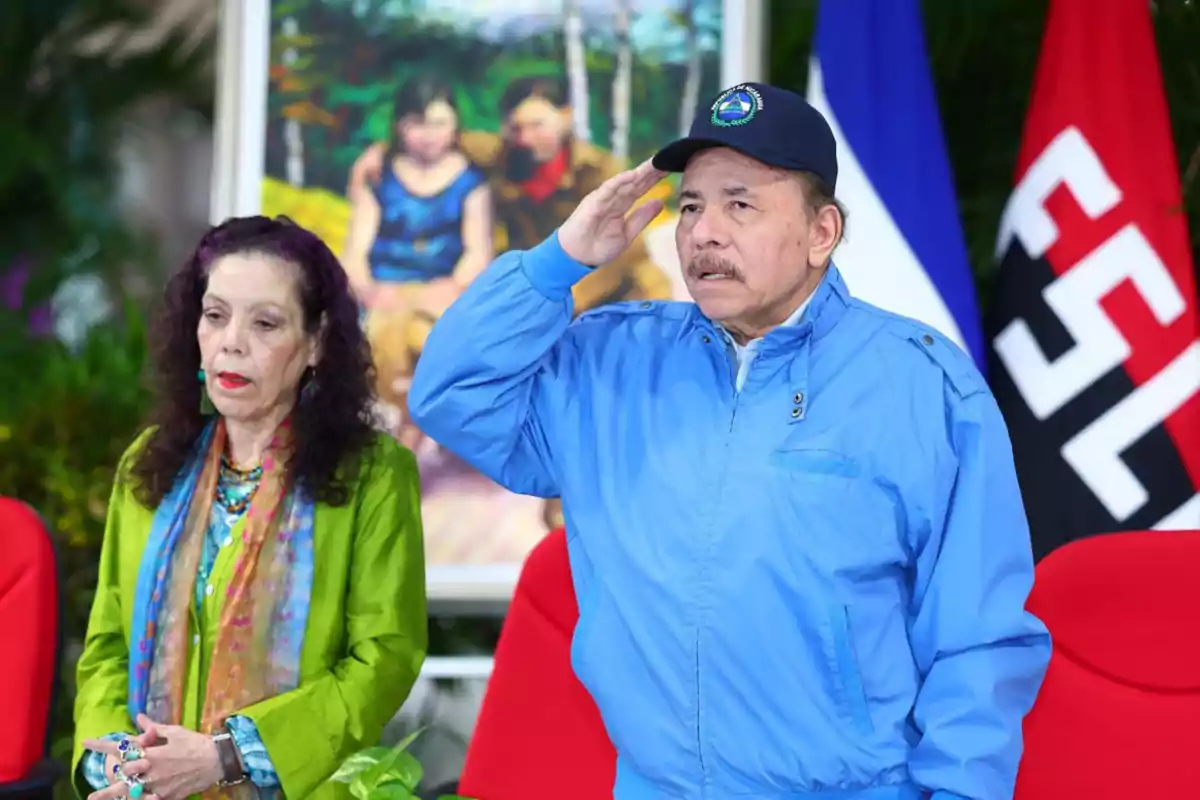 Dos personas están de pie frente a un fondo con banderas y una pintura, una de ellas lleva una chaqueta azul y gorra mientras hace un saludo.