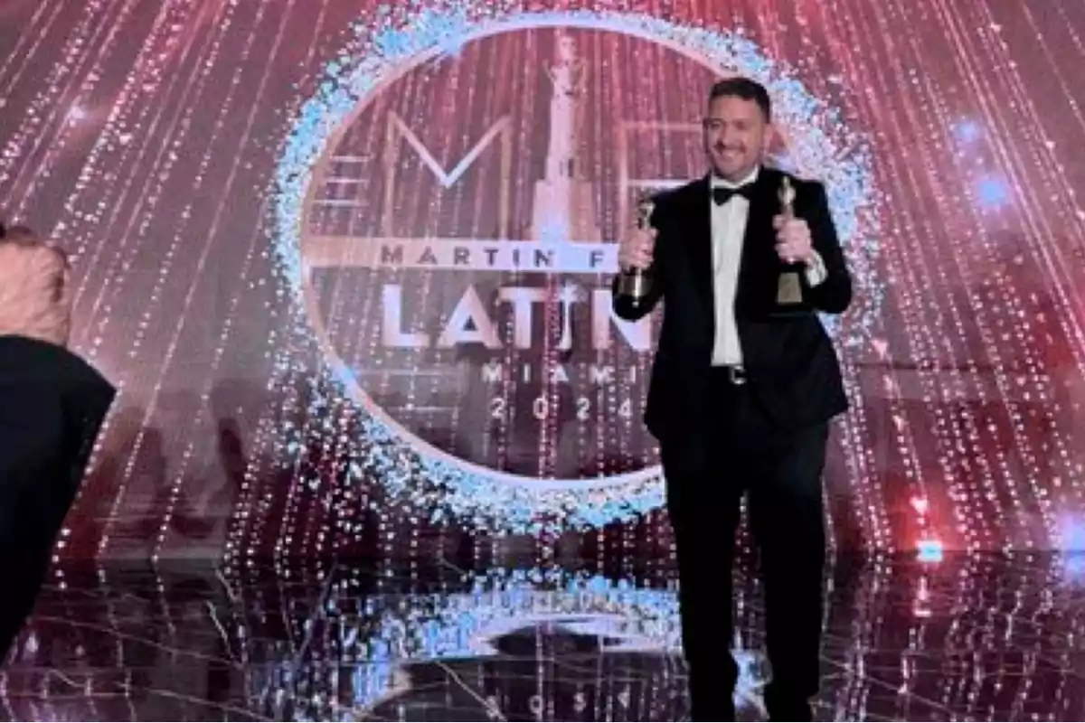 Persona en traje sosteniendo dos premios en un escenario decorado con luces y un fondo que dice "Martín Fierro Latino Miami 2024".