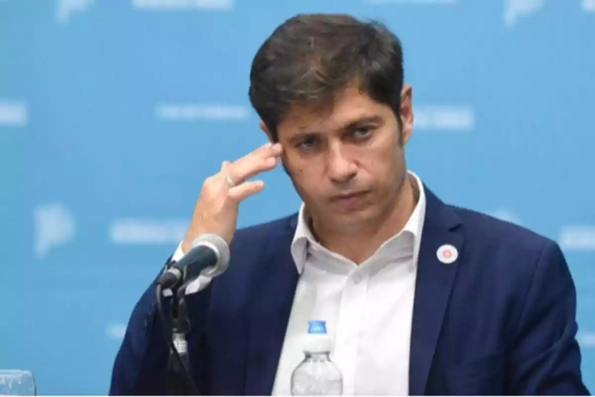 Hombre en traje oscuro con camisa blanca frente a un micrófono y una botella de agua sobre una mesa con fondo azul.