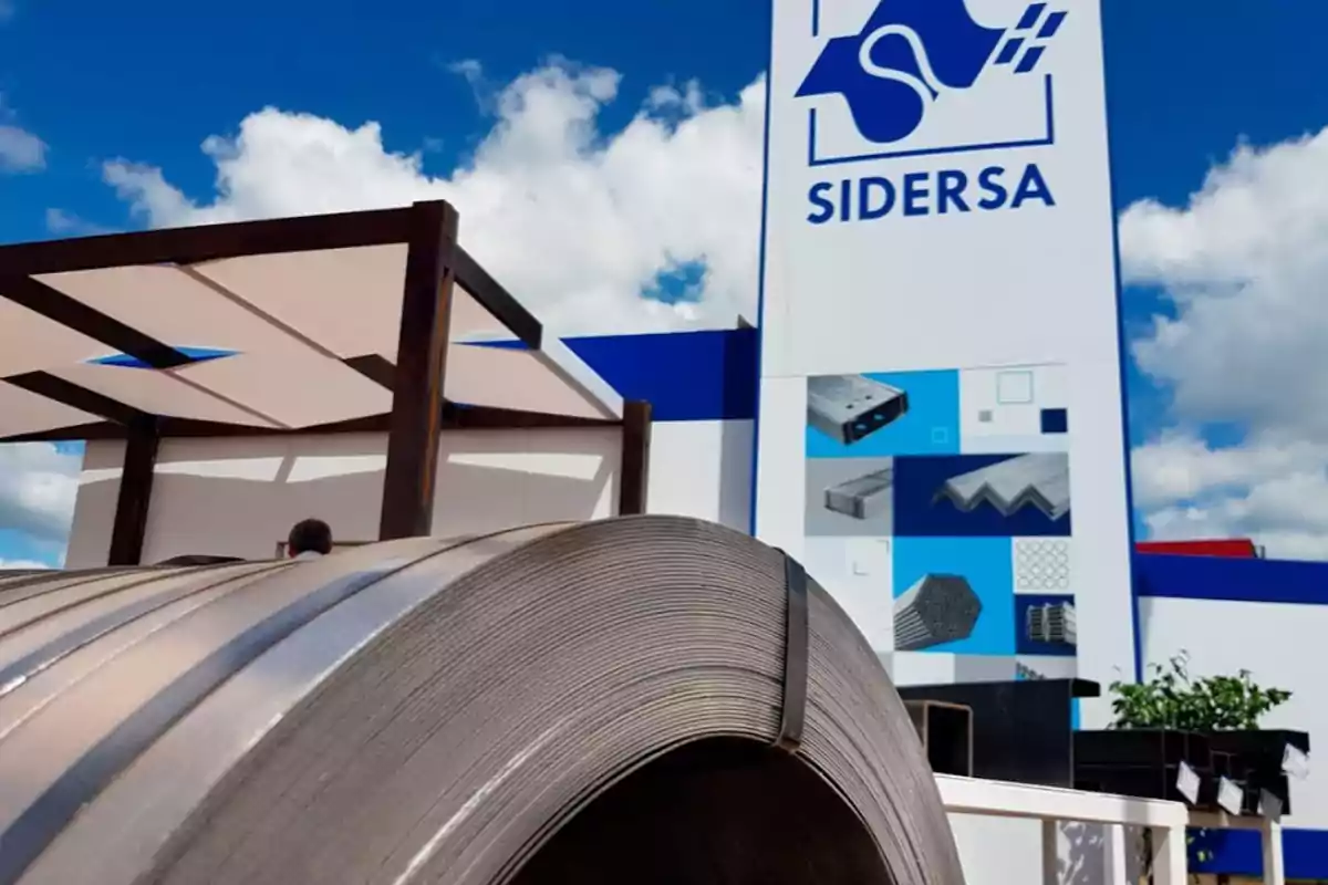 Rollo de metal frente a un edificio con el logo de Sidersa y un cielo azul con nubes.