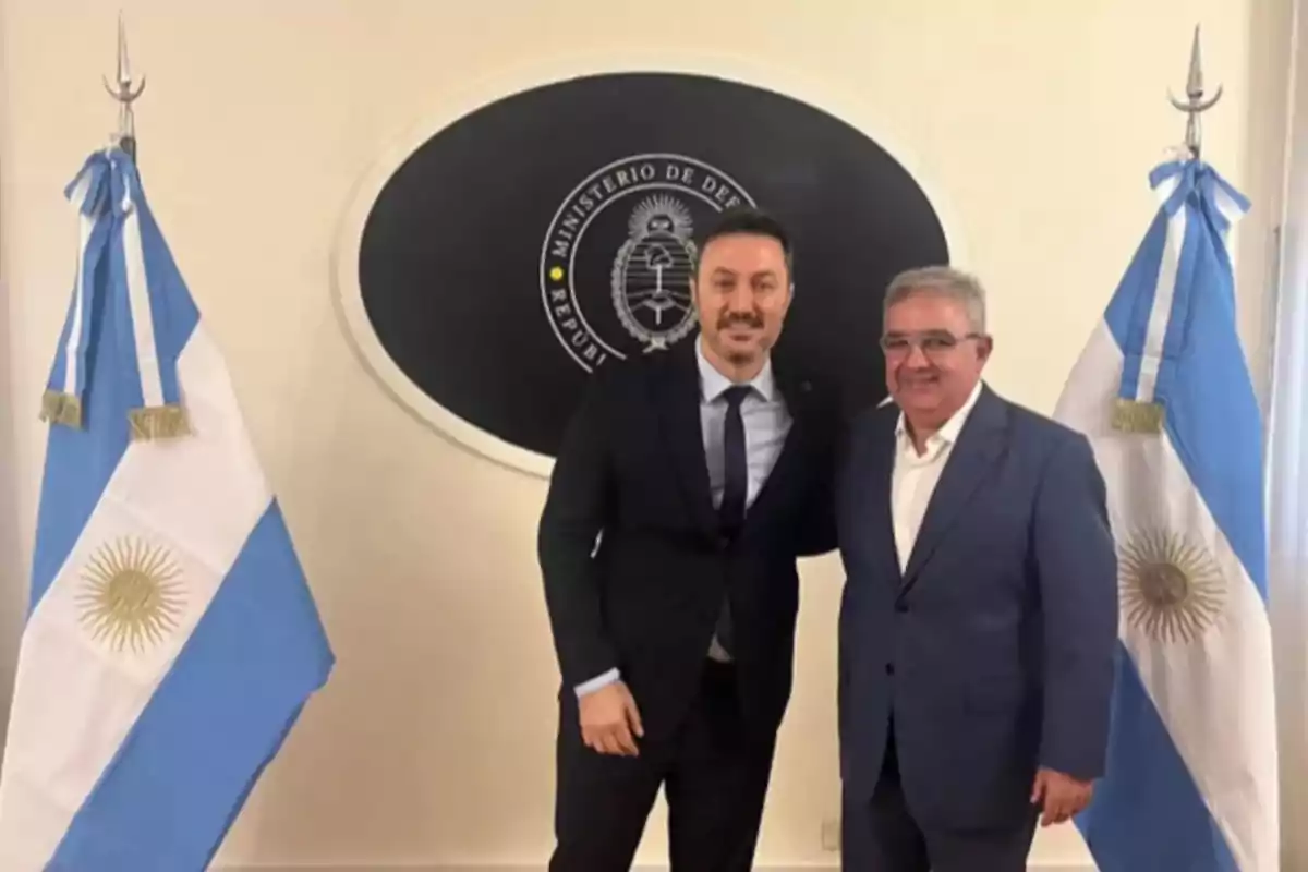 Dos hombres de traje posan frente a un emblema del Ministerio de Defensa de Argentina, flanqueados por banderas argentinas.