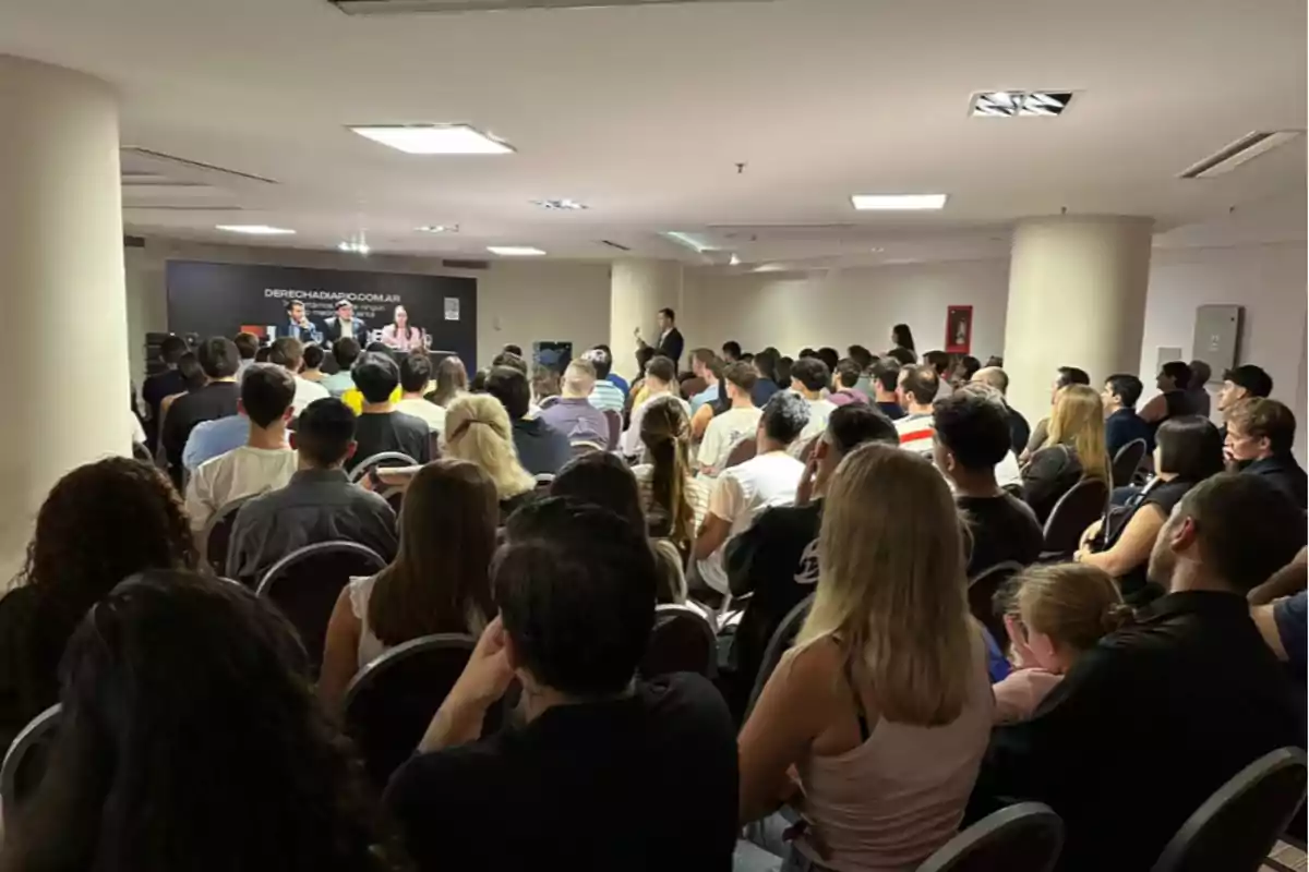 Una sala llena de personas asistiendo a una conferencia con un panel de oradores al frente.