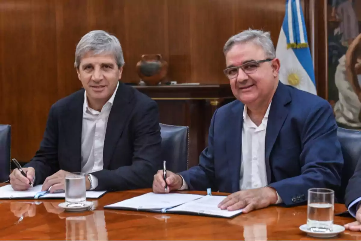 Dos hombres sentados en una mesa de madera firmando documentos con una bandera de Argentina en el fondo.