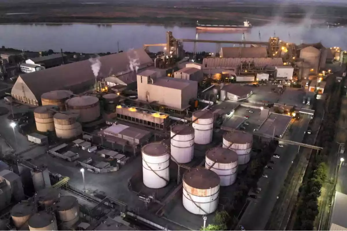 Vista aérea de una planta industrial con varios tanques de almacenamiento y edificios, ubicada cerca de un río.