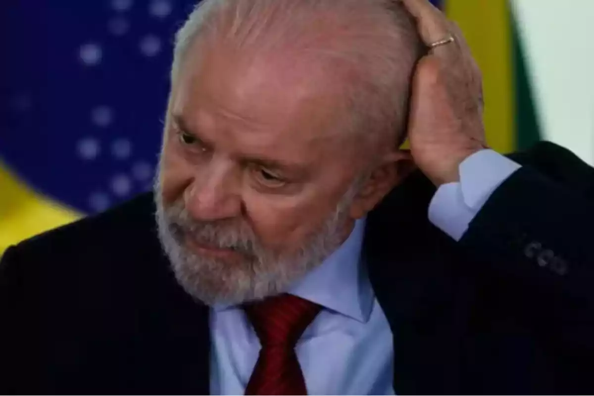 Un hombre con barba y traje oscuro se toca la cabeza con una mano frente a una bandera.