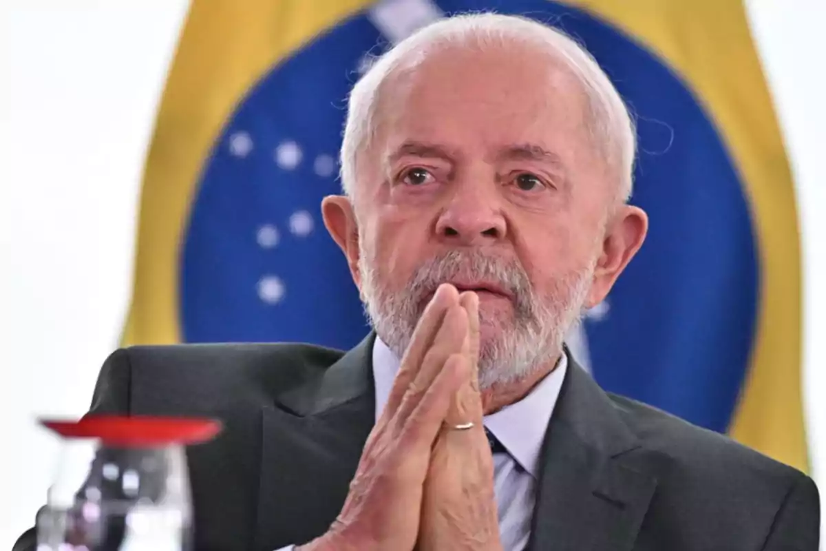 Un hombre mayor con barba y traje está con las manos juntas frente a una bandera de Brasil.