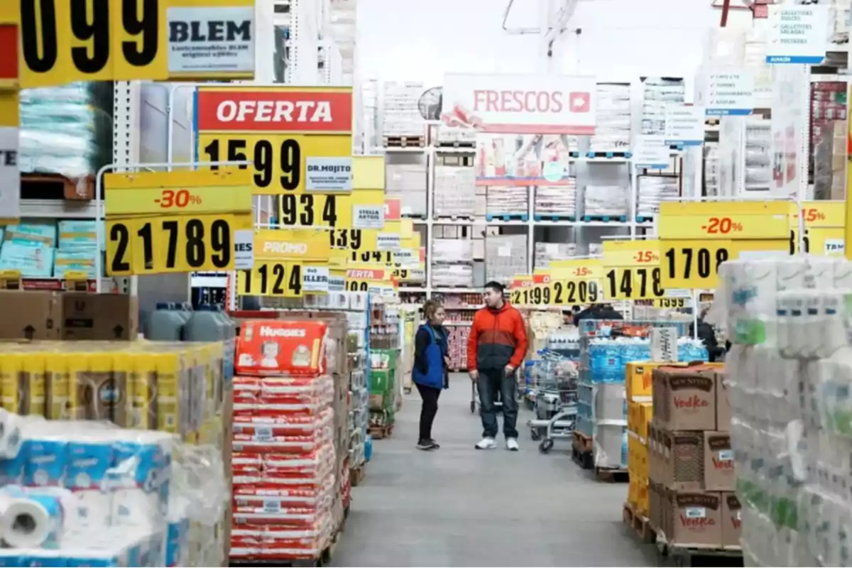 Pasillo de un supermercado mayorista con estantes llenos de productos y carteles de ofertas y precios destacados.