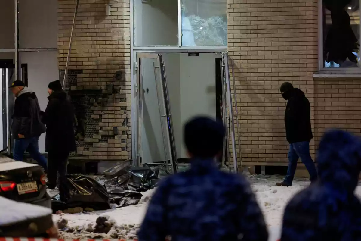 Personas caminando cerca de un edificio con daños en la fachada y nieve en el suelo.