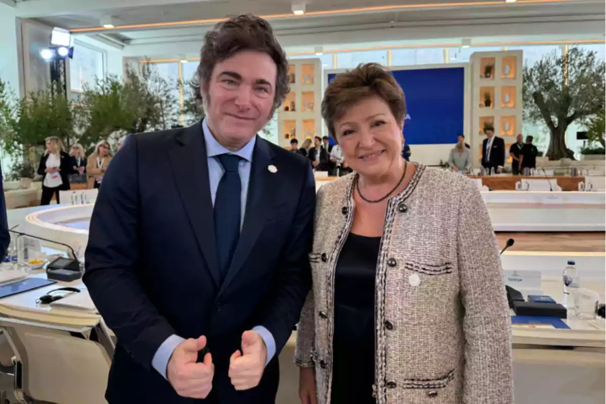 Dos personas posan juntas en un entorno de conferencia con mesas y sillas blancas y árboles decorativos al fondo.