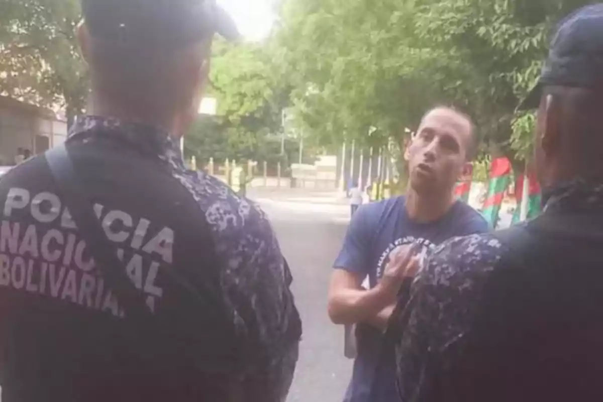 Un hombre de pie frente a dos oficiales de la Policía Nacional Bolivariana en una calle con árboles y banderas al fondo.