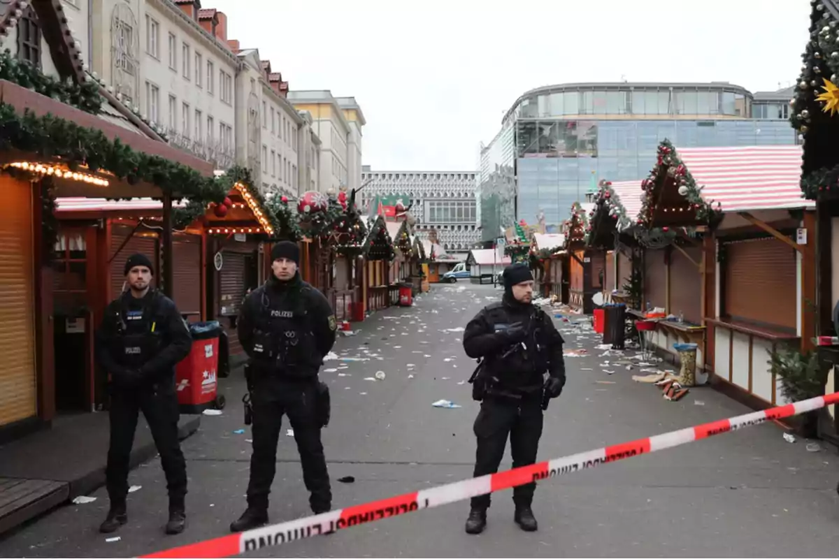 Tres policías de pie frente a un mercado navideño cerrado con cintas de seguridad y decoraciones festivas en los puestos.