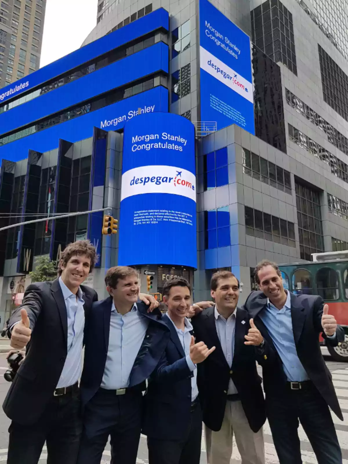 Un grupo de cinco hombres sonrientes con trajes posan frente a un edificio con pantallas azules que muestran mensajes de felicitación de Morgan Stanley a Despegar.com en una ciudad urbana.