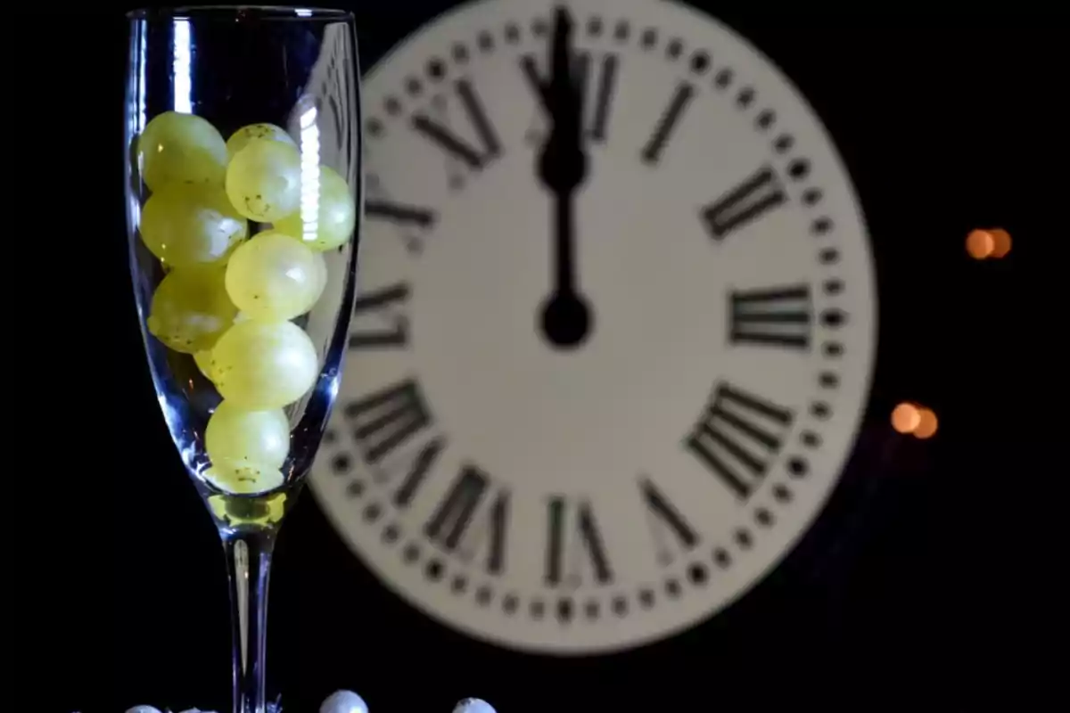 Una copa de champán con uvas frente a un reloj marcando la medianoche.