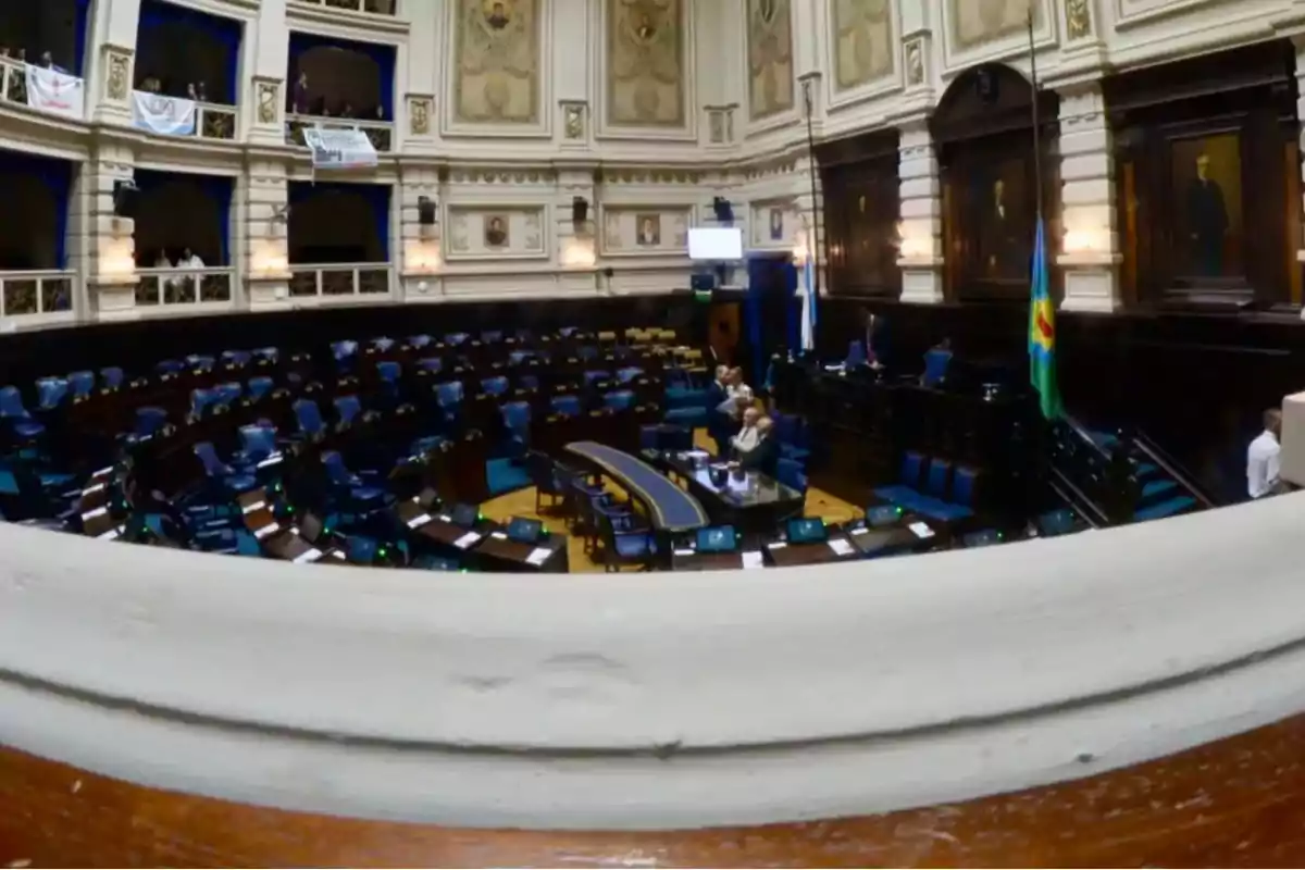 Una sala de asambleas vacía con sillas azules y paredes decoradas con retratos y molduras.