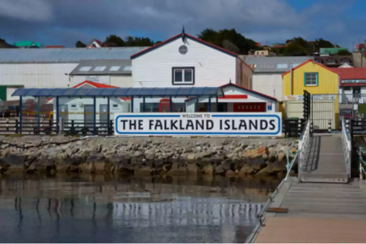 Un cartel de bienvenida a las Islas Malvinas frente a un muelle con edificios coloridos al fondo.